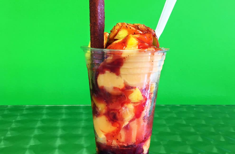 Against a green background, a plastic parfait cup is filled with scoops of mango ice cream with reddish brown chili syrup, a white spoon, and a tamarind candy stick