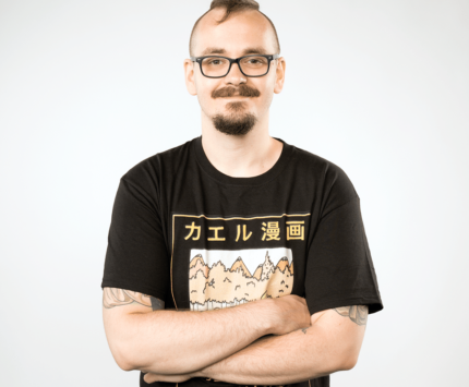 A man dressed in a black t-shirt and black pants, with black glasses, standing with his arms crossed across his chest