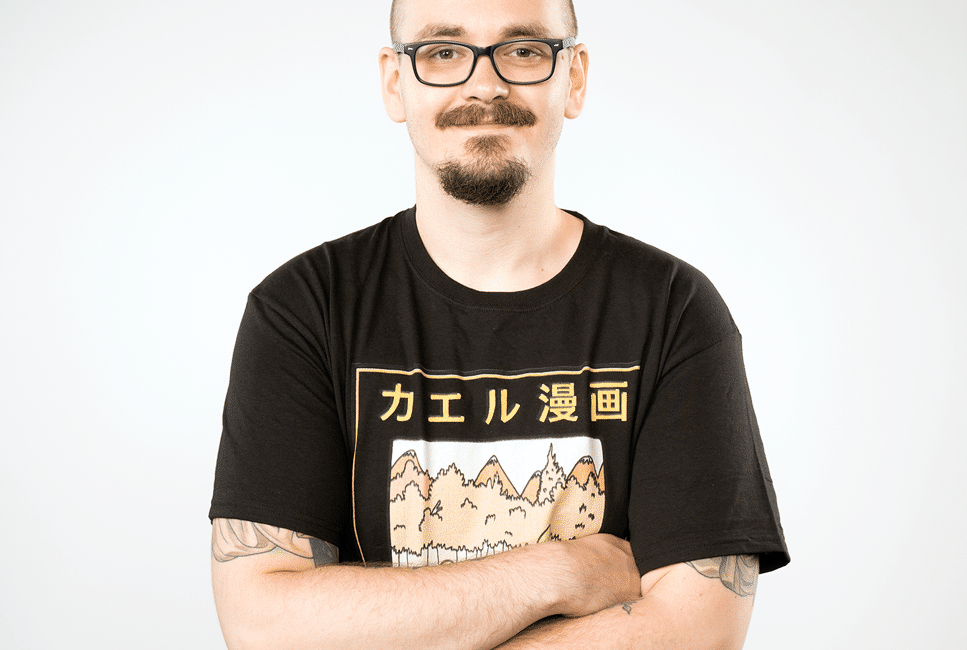 A man dressed in a black t-shirt and black pants, with black glasses, standing with his arms crossed across his chest