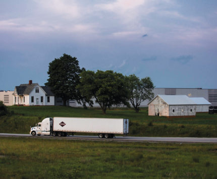 A semi truck drives past a farm house with a warehouse behind it