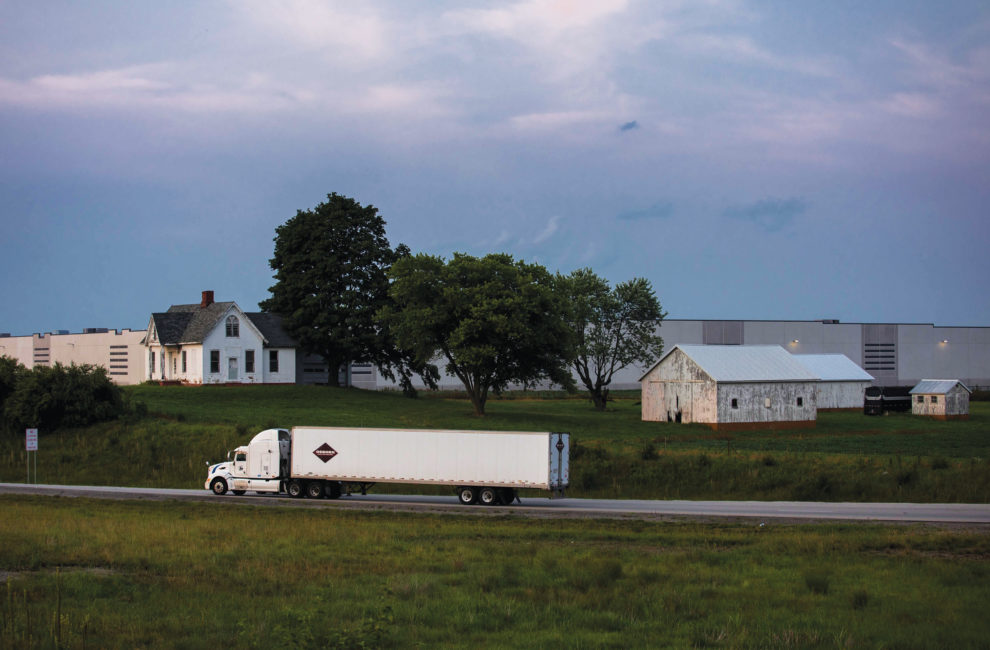 A semi truck drives past a farm house with a warehouse behind it