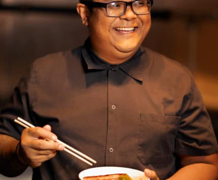 Man in a baseball cap and brown shirt holding a white bowl and chopsticks