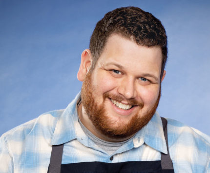 Man in a blue plaid shirt and dark apron, with