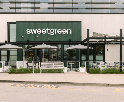 an exterior view of Sweetgreen restaurant, with a green sign, white seating, and umbrellas