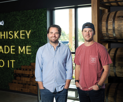West Fork Whiskey and The Mash House co-owners blake jones and David McIntyre stand in front of a neon sign saying "(local) whiskey made me do it"